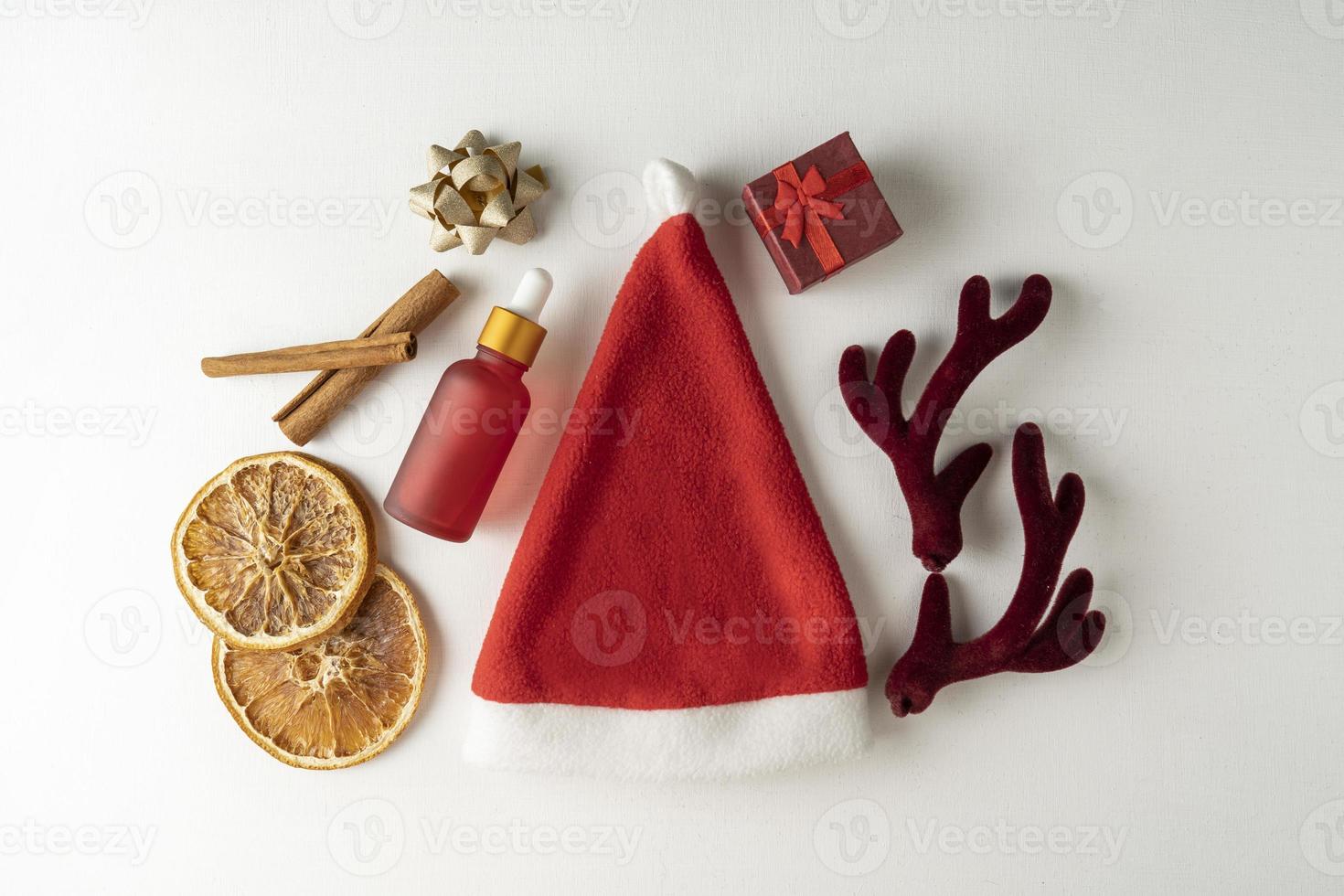 A face serum or essential oil for Christmas holidays in a red dropper bottle lying on a white background with golden bows, cinnamon and dried oranges around photo