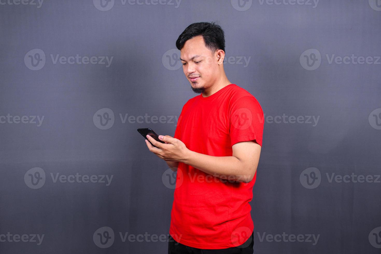 Portrait of a young cheerful asian man wearing red t-shirt standing isolated over gray background, using mobile phone photo