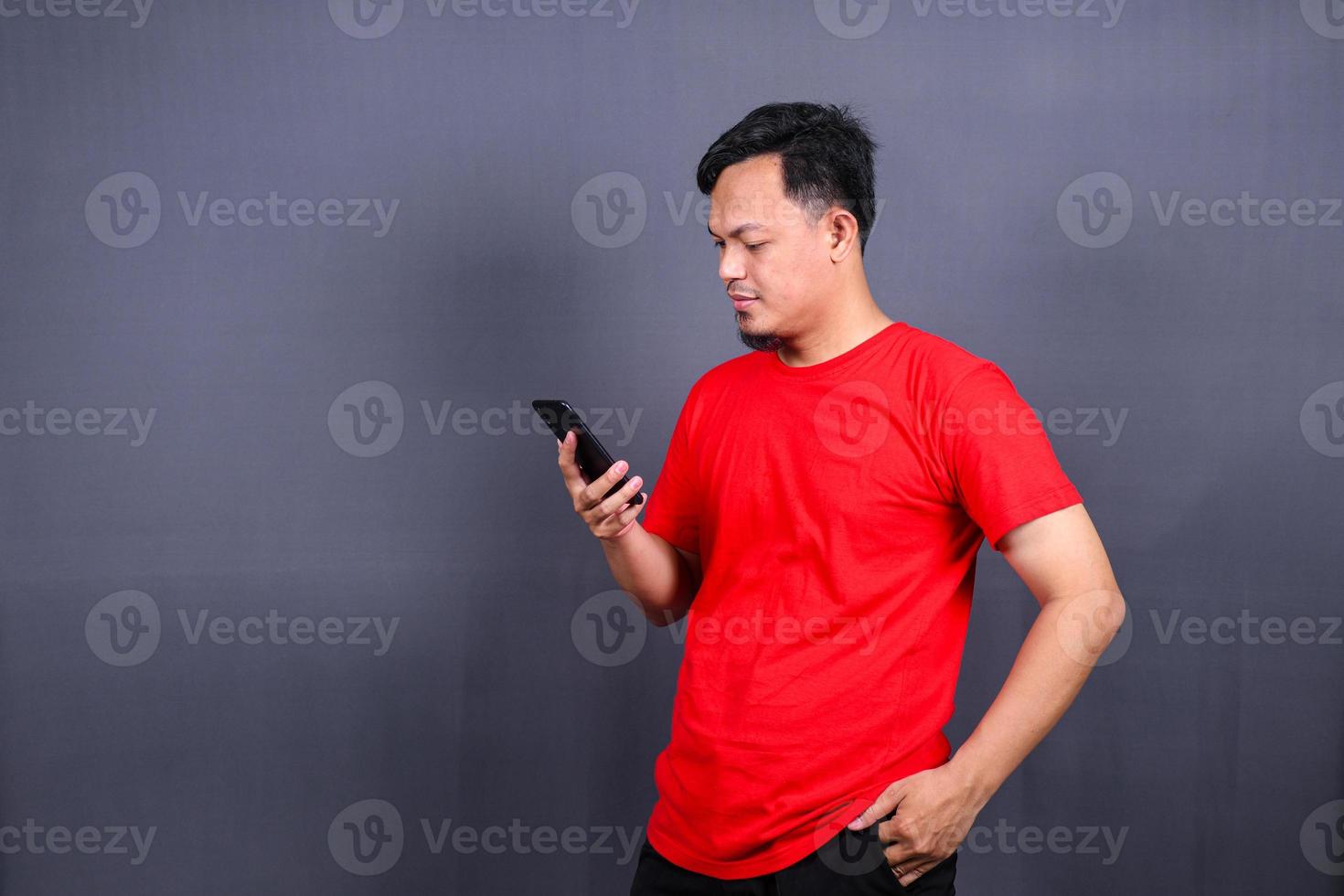 Portrait of a young cheerful asian man wearing red t-shirt standing isolated over gray background, using mobile phone photo