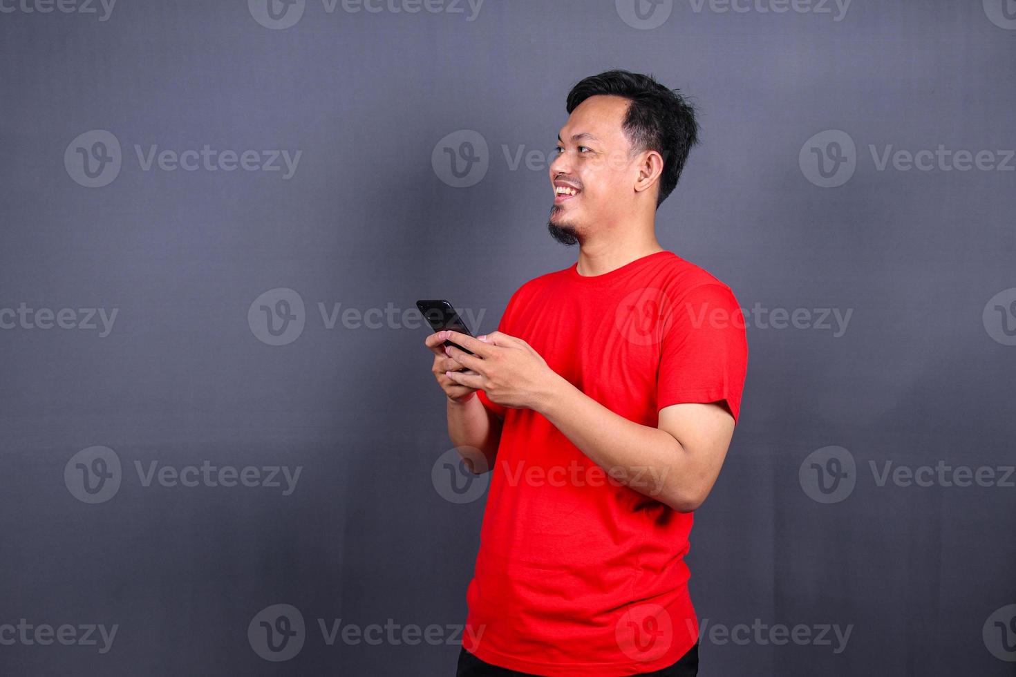 Portrait of a young cheerful asian man wearing red t-shirt standing isolated over gray background, using mobile phone photo