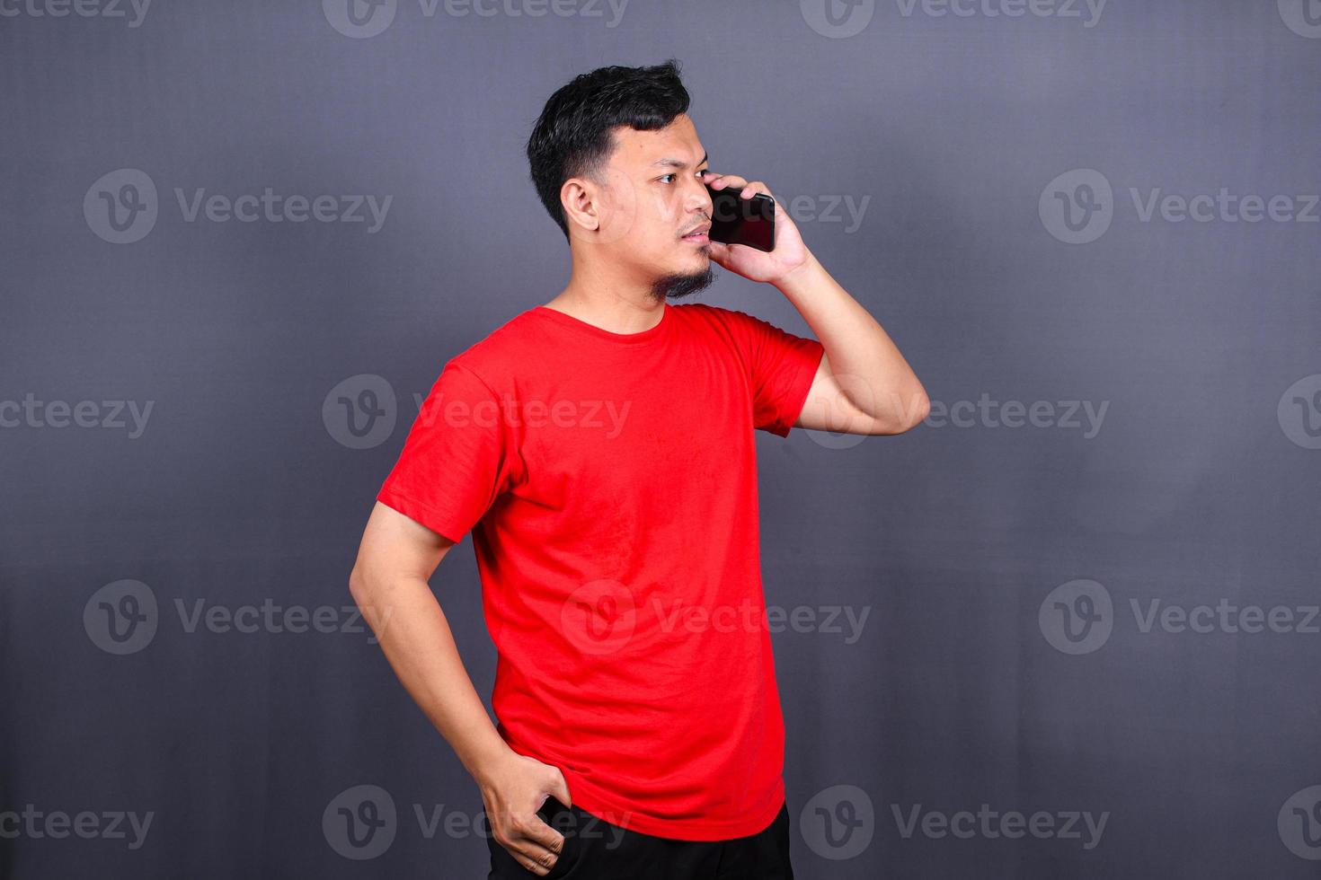 Portrait of a young cheerful asian man wearing red t-shirt standing isolated over gray background, using mobile phone photo