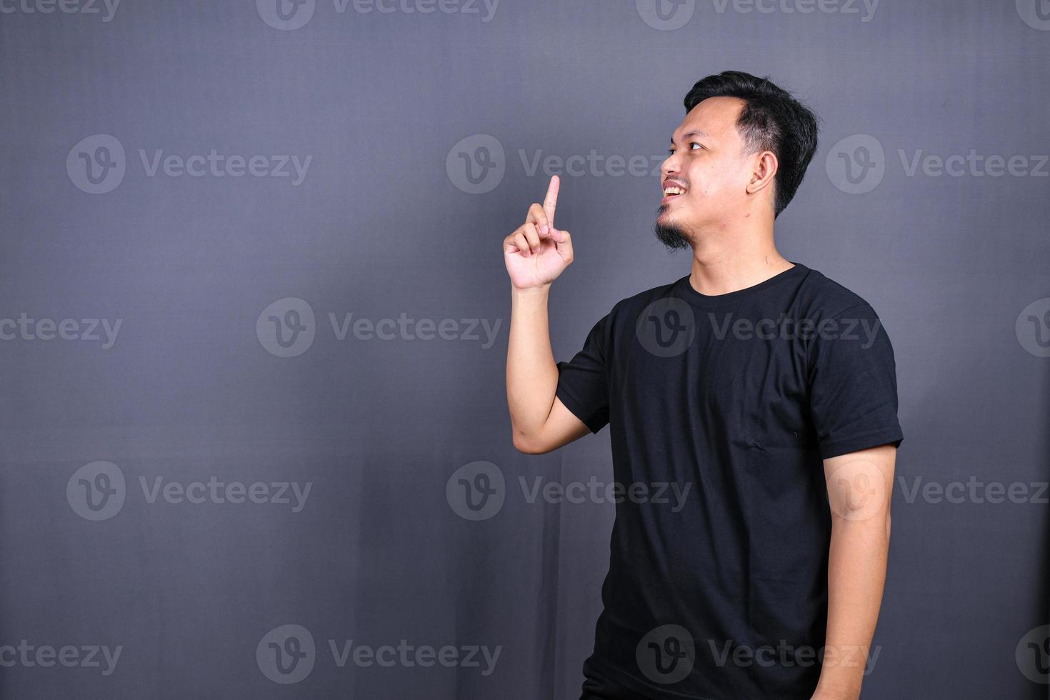 Portrait of a happy young asian man in black t-shirt pointing fingers away at copy space isolated over gray background photo