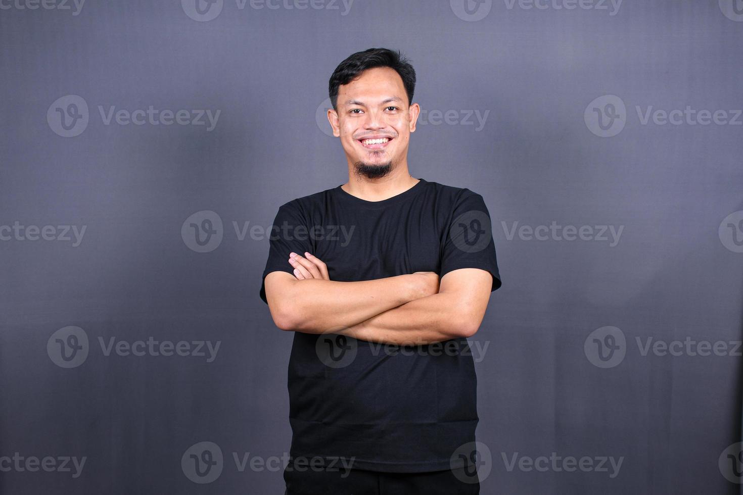 un hombre asiático sonriente y apuesto con una camiseta negra de pie con los brazos cruzados aislado en un fondo gris foto