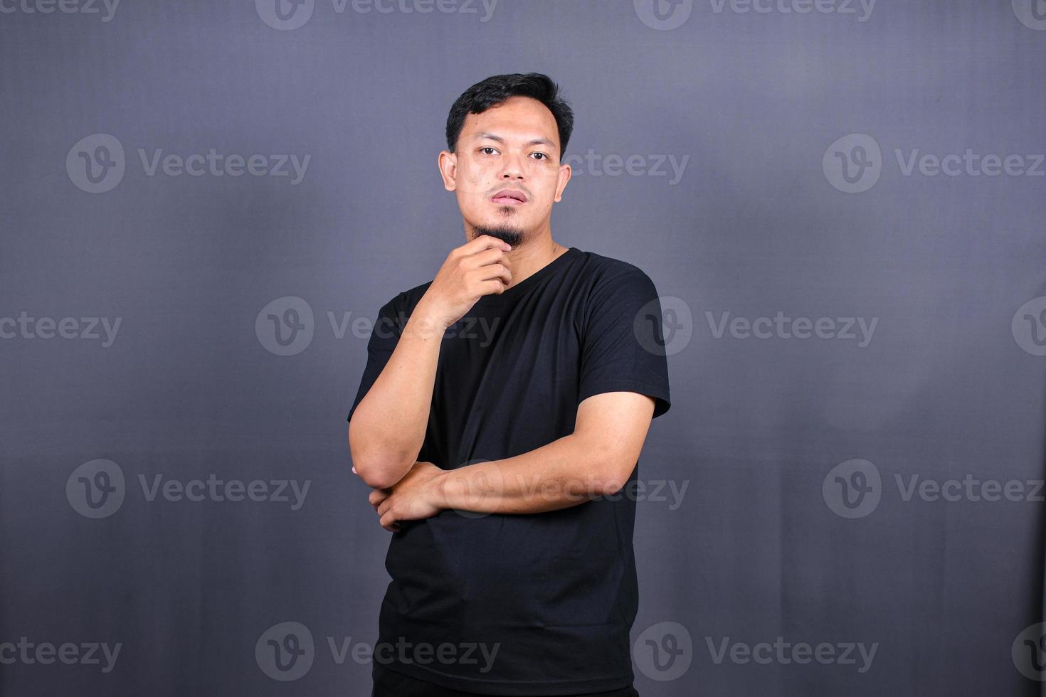 young Asian man holds chin and looks offended at camera feels angry awakes in bad mood isolated over gray background photo