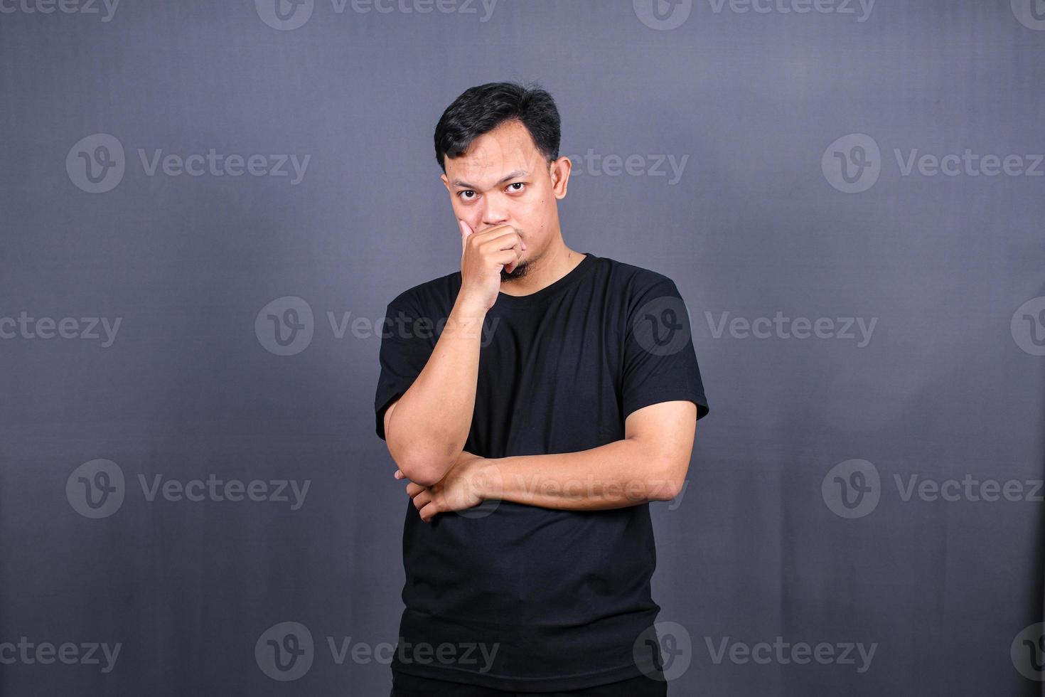 young Asian man holds chin and looks offended at camera feels angry awakes in bad mood isolated over gray background photo