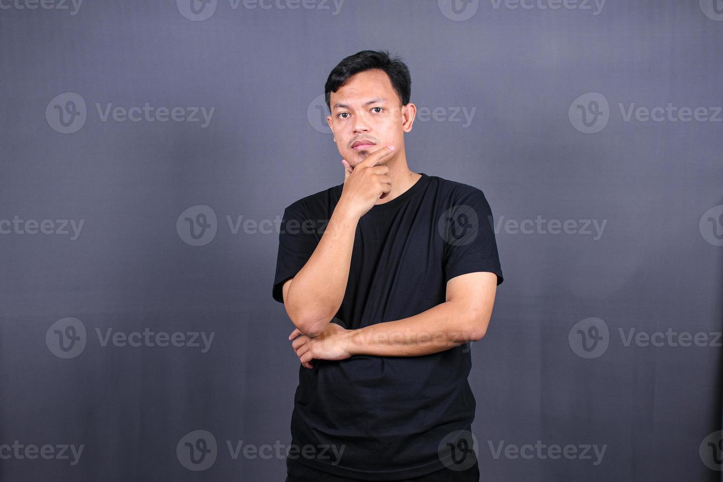 young Asian man holds chin and looks offended at camera feels angry awakes in bad mood isolated over gray background photo