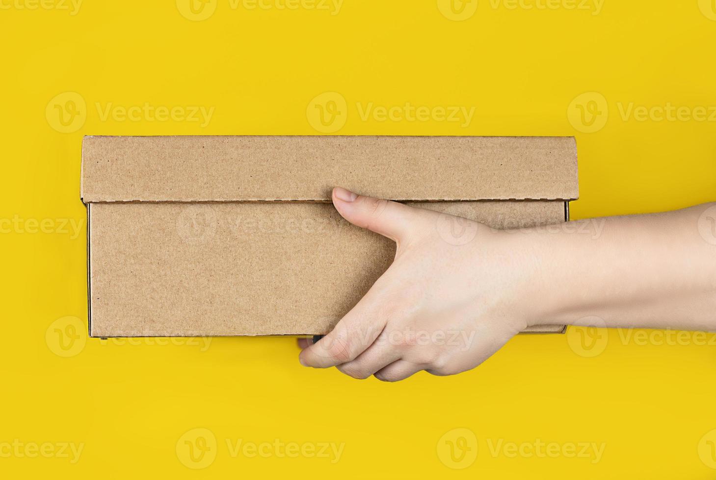 Large cardboard box in hands on a trendy yellow background. Horizontal photo. Concept - delivery of orders to your home by courier, receipt of parcels, safe distance photo