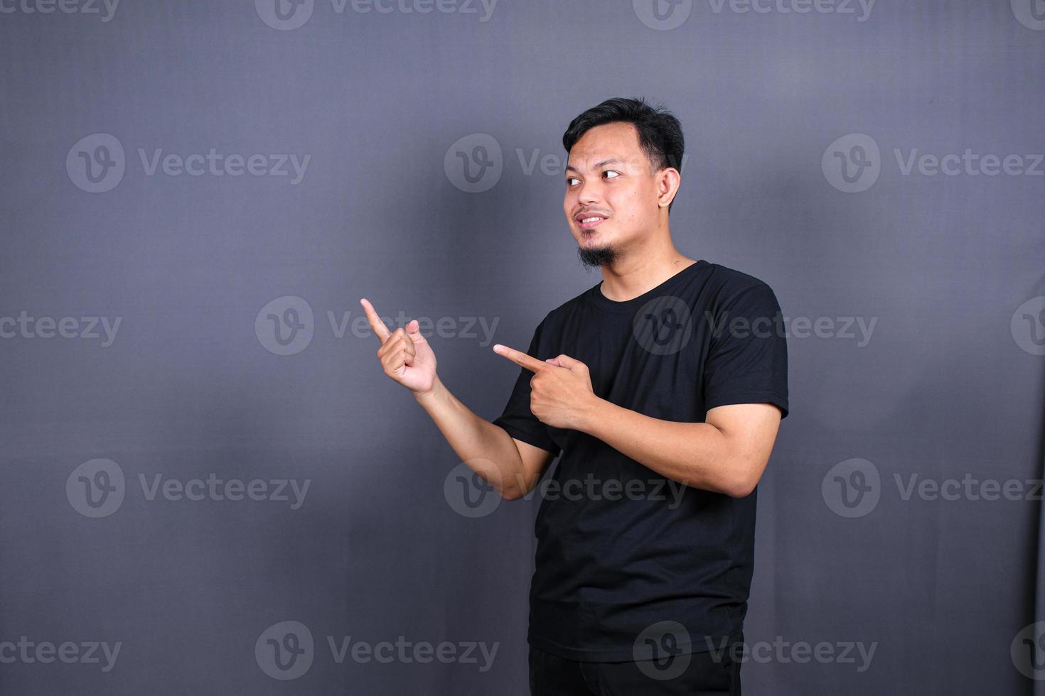 retrato de un joven asiático feliz con una camiseta negra señalando con el dedo el espacio de copia aislado sobre fondo gris foto