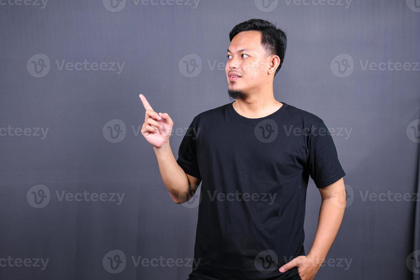 Portrait of a happy young asian man in black t-shirt pointing fingers away at copy space isolated over gray background photo