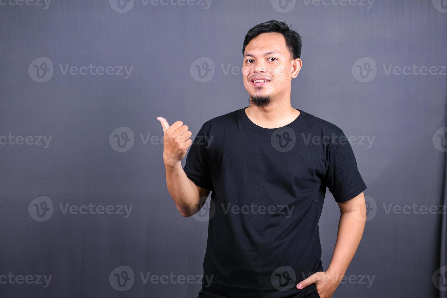 retrato de un joven asiático feliz con una camiseta negra señalando con el dedo el espacio de copia aislado sobre fondo gris foto