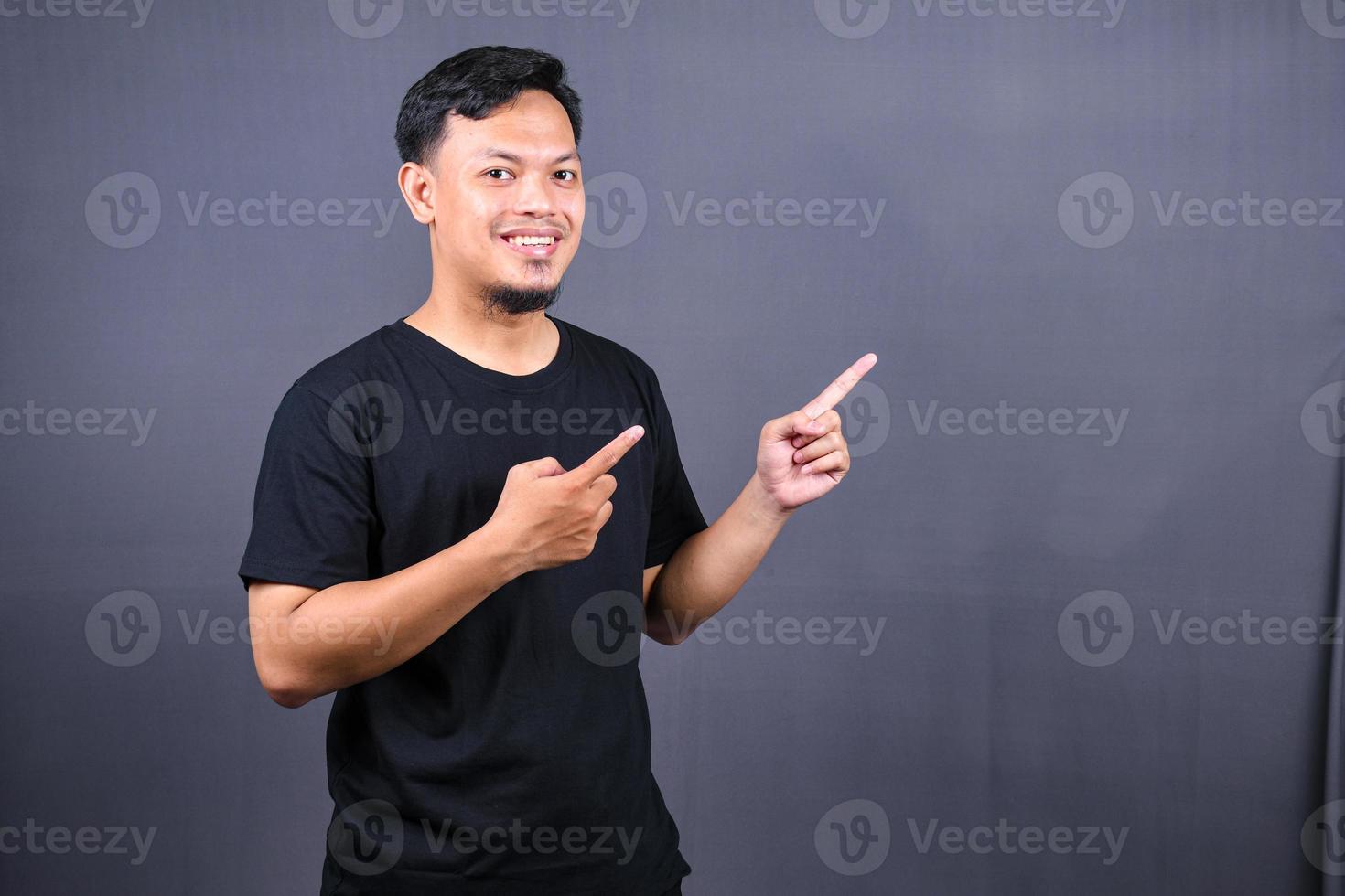 Portrait of a happy young asian man in black t-shirt pointing fingers away at copy space isolated over gray background photo