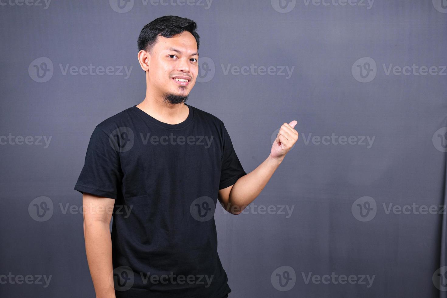 retrato de un joven asiático feliz con una camiseta negra señalando con el dedo el espacio de copia aislado sobre fondo gris foto