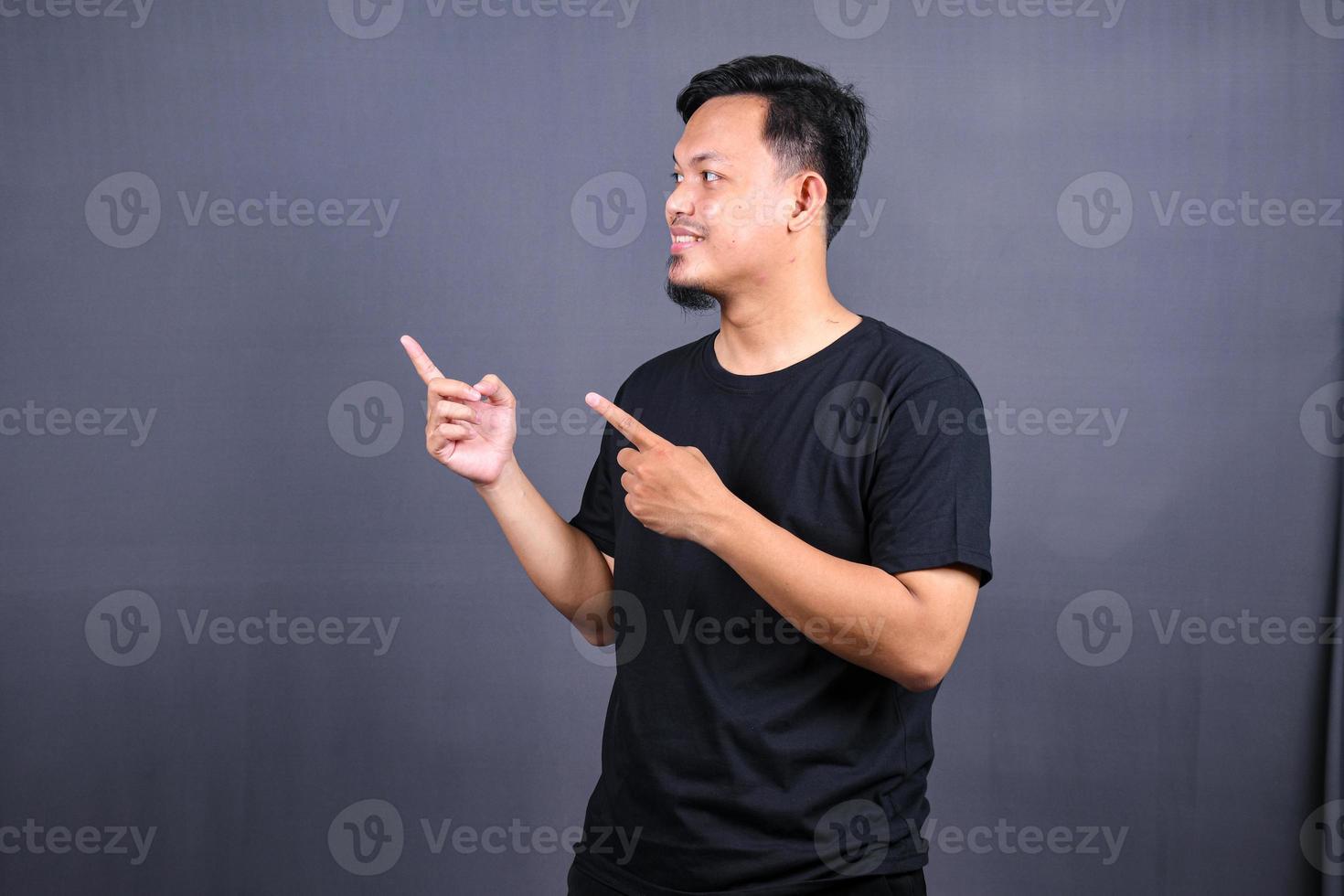Portrait of a happy young asian man in black t-shirt pointing fingers away at copy space isolated over gray background photo