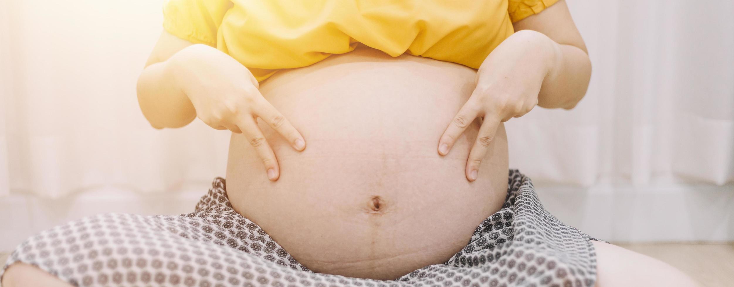 Young adult pregnant woman in white clothes touching big naked belly with hands. Showing shape. Pregnancy concept. Expectation time. Closeup. Isolated on light gray wall background. photo