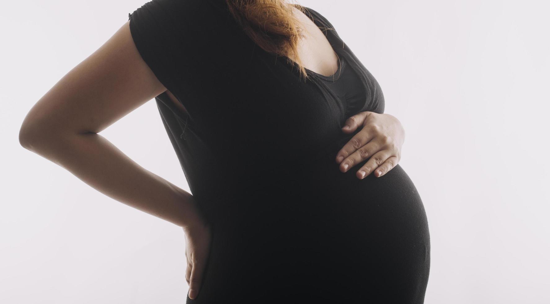 Young adult pregnant woman in white clothes touching big naked belly with hands. Showing shape. Pregnancy concept. Expectation time. Closeup. Isolated on light gray wall background. photo