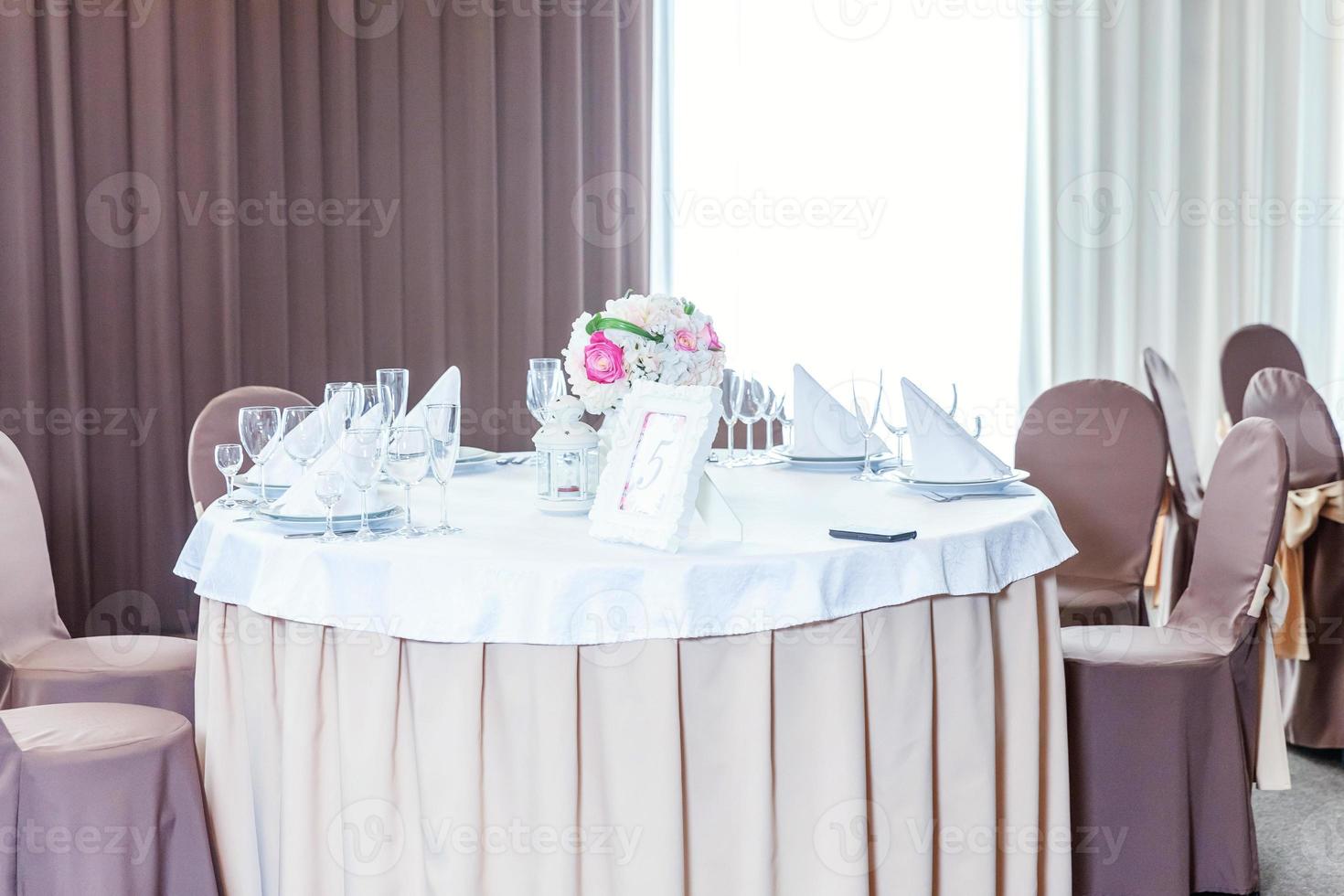 Fancy table set for dinner with flower composition in restaurant, luxury interior background. Wedding elegant banquet decoration and items for food arranged by catering service on white table. photo