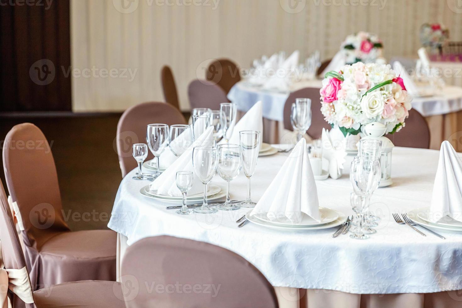 Fancy table set for dinner with flower composition in restaurant, luxury interior background. Wedding elegant banquet decoration and items for food arranged by catering service on white table. photo