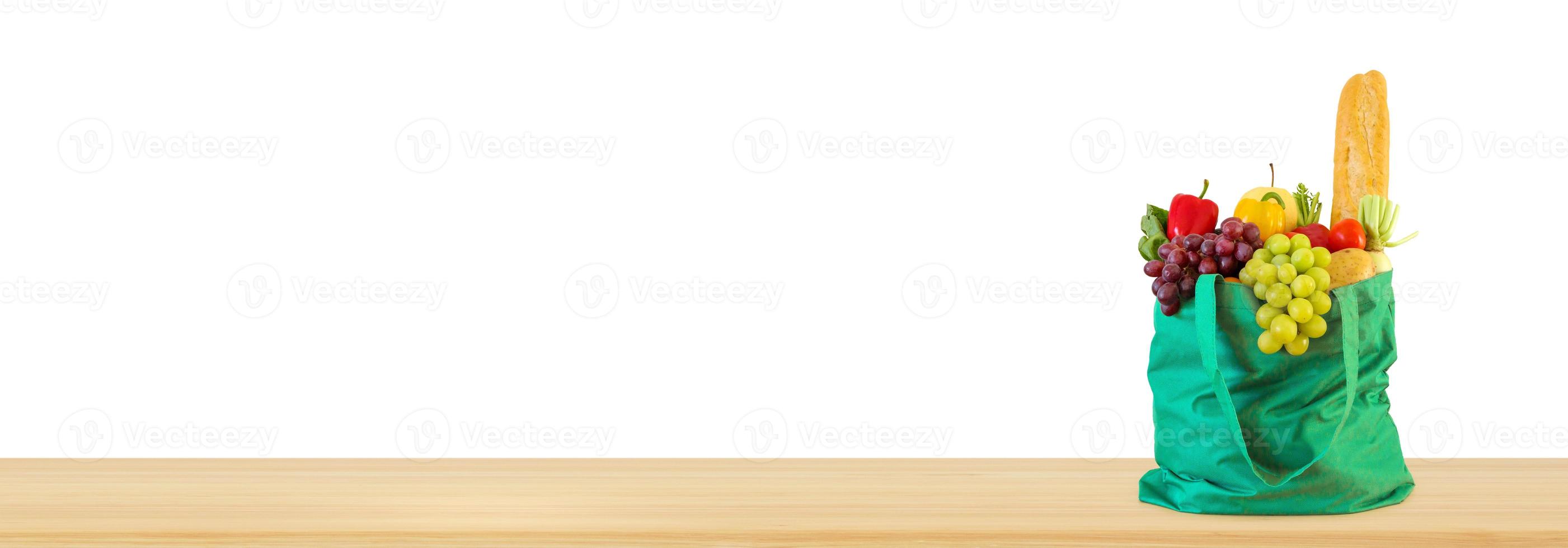 Fresh fruits and vegetables grocery product in green reusable shopping bag on wood table top isolated on white background photo