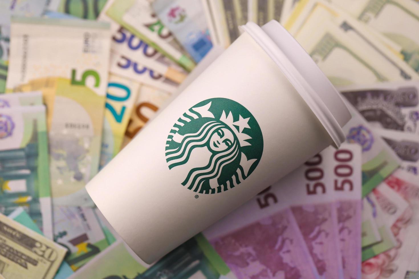 KHARKIV, UKRAINE - DECEMBER 16, 2021 White paper cup with Starbucks logo and money bills. Starbucks is the world's largest coffee house with over 20,000 stores. photo