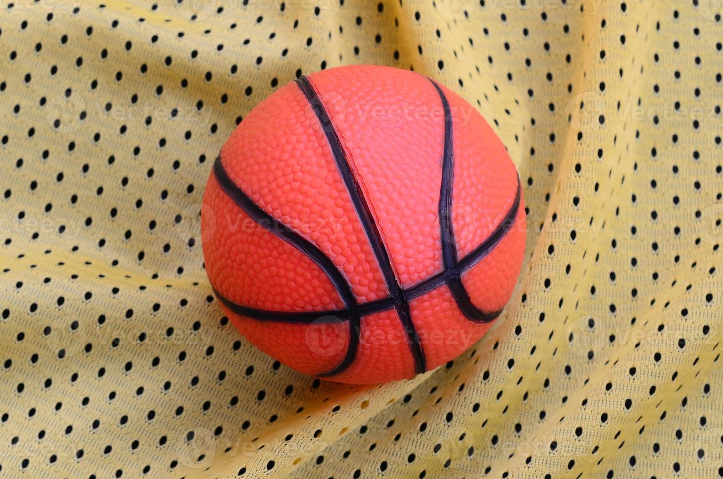 Small orange rubber basketball lies on a yellow sport jersey clothing fabric texture and background with many folds photo