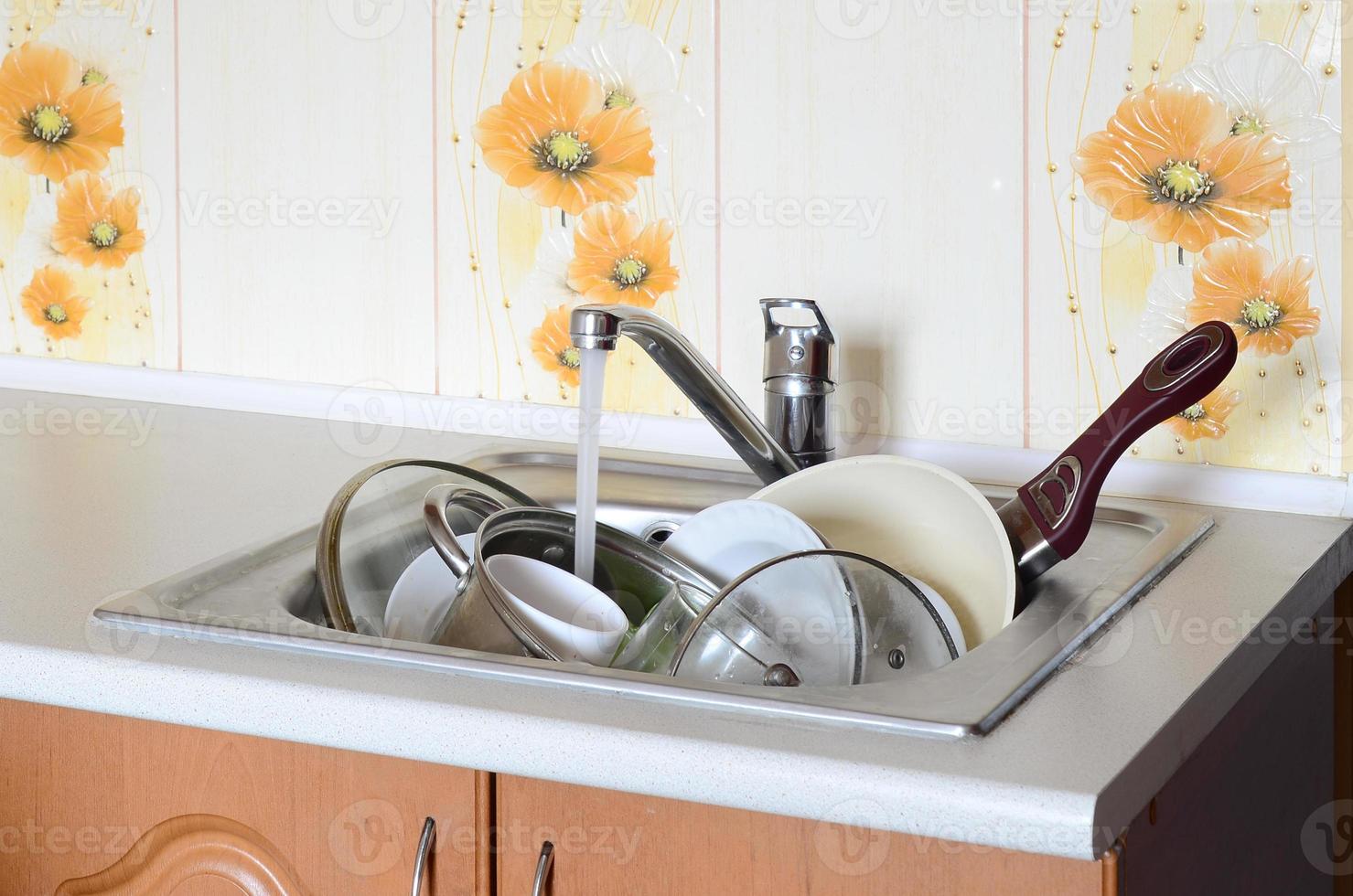 Dirty dishes and unwashed kitchen appliances lie in foam water under a tap from a kitchen faucet photo