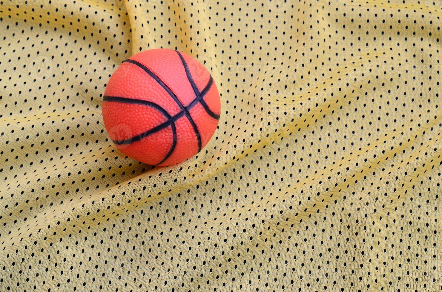 Small orange rubber basketball lies on a yellow sport jersey clothing fabric texture and background with many folds photo