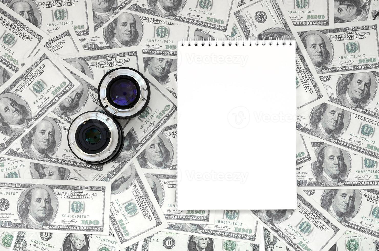 Two photographic lenses and white notebook lie on the background of a lot of dollar bills. Space for text photo