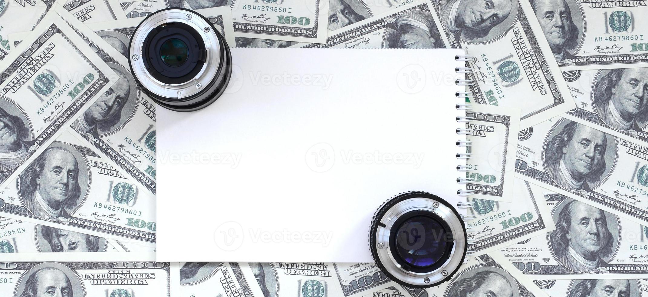 Two photographic lenses and white notebook lie on the background of a lot of dollar bills. Space for text photo