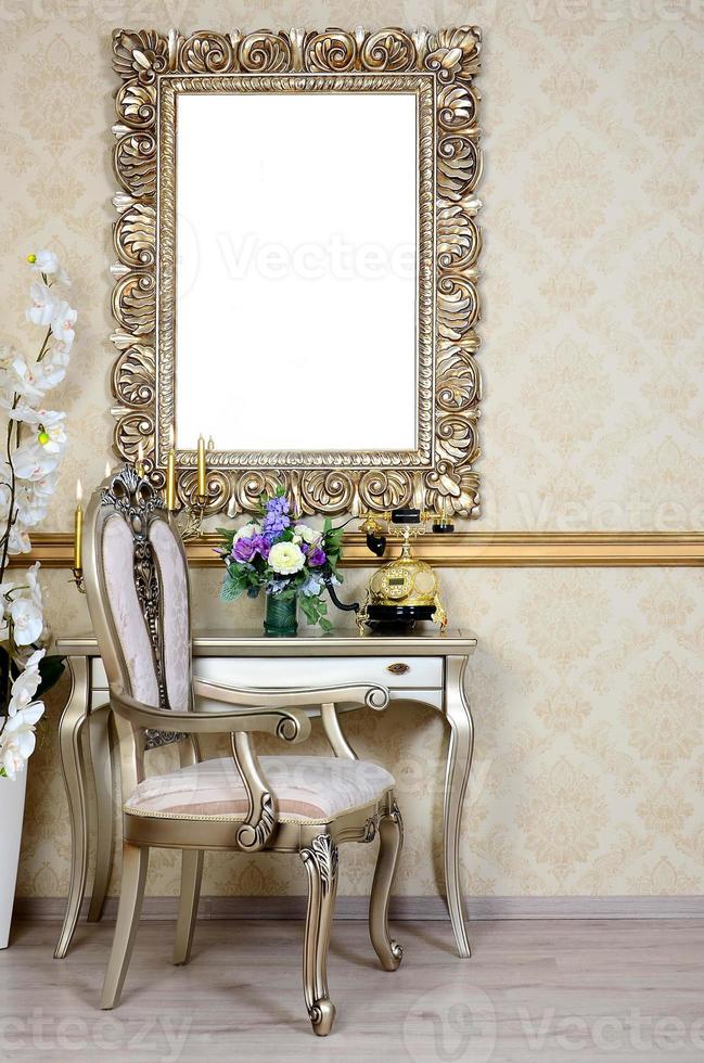 A fragment of a retro interior with a chair and table, on which is a telephone and a vase of flowers. Above the table hangs an empty picture frame for your photo
