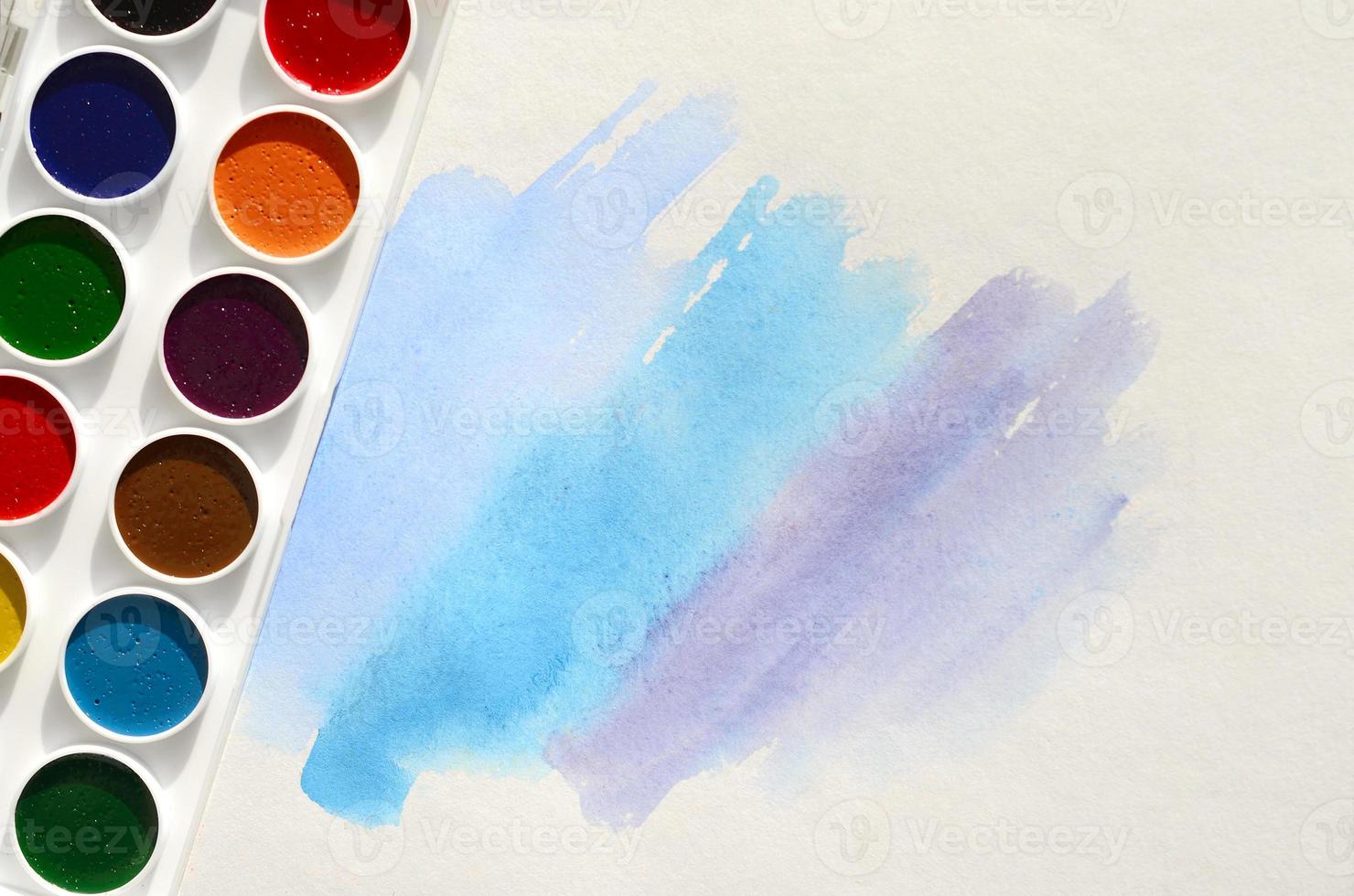A new set of watercolors lies on a sheet of paper, which shows an abstract watercolor drawing in the form of blue strokes. The concept of amateur painting among teenagers photo