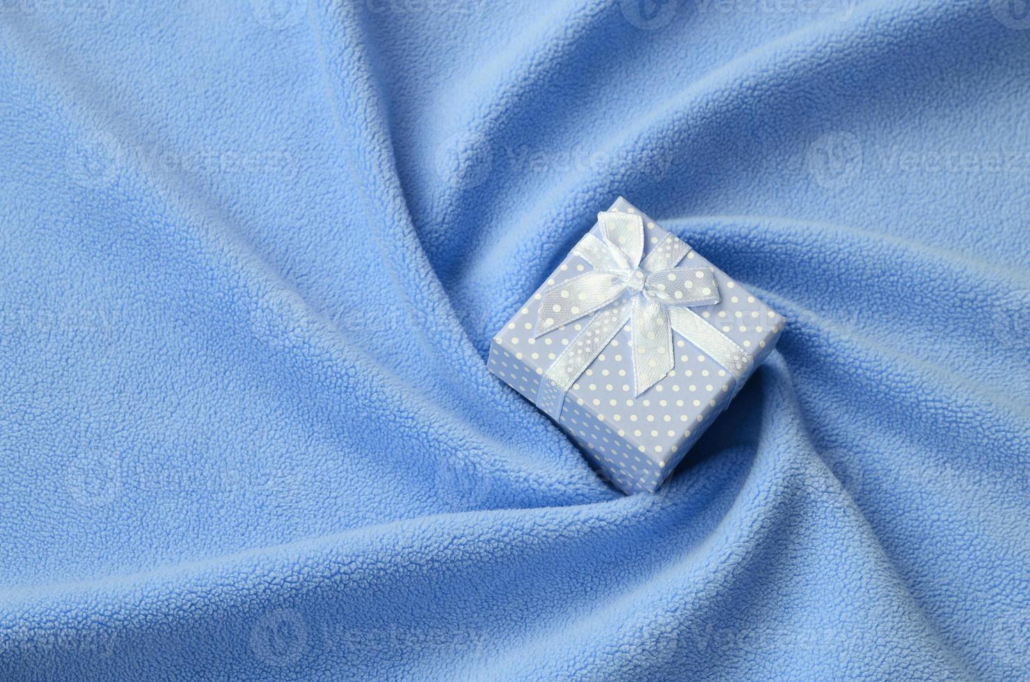 A small gift box in blue with a small bow lies on a blanket of soft and furry light blue fleece fabric with a lot of relief folds. Packing for a gift to your lovely girlfriend photo