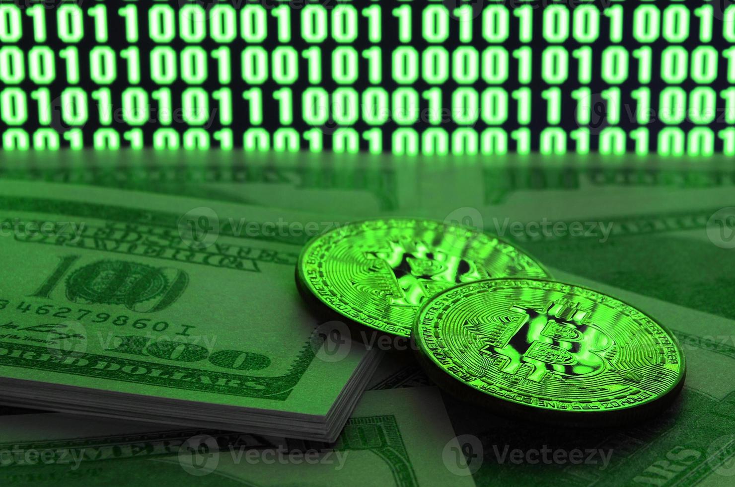 Two bitcoins lies on a pile of dollar bills on the background of a monitor depicting a binary code of bright green zeros and one units on a black background. Low key lighting photo