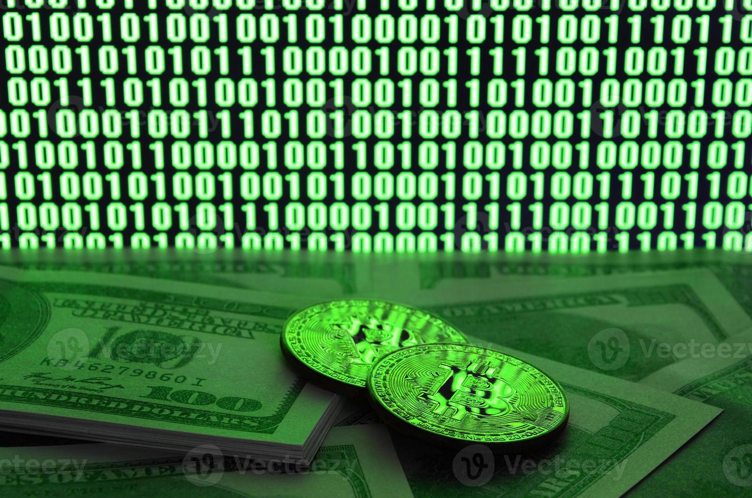 Two bitcoins lies on a pile of dollar bills on the background of a monitor depicting a binary code of bright green zeros and one units on a black background. Low key lighting photo