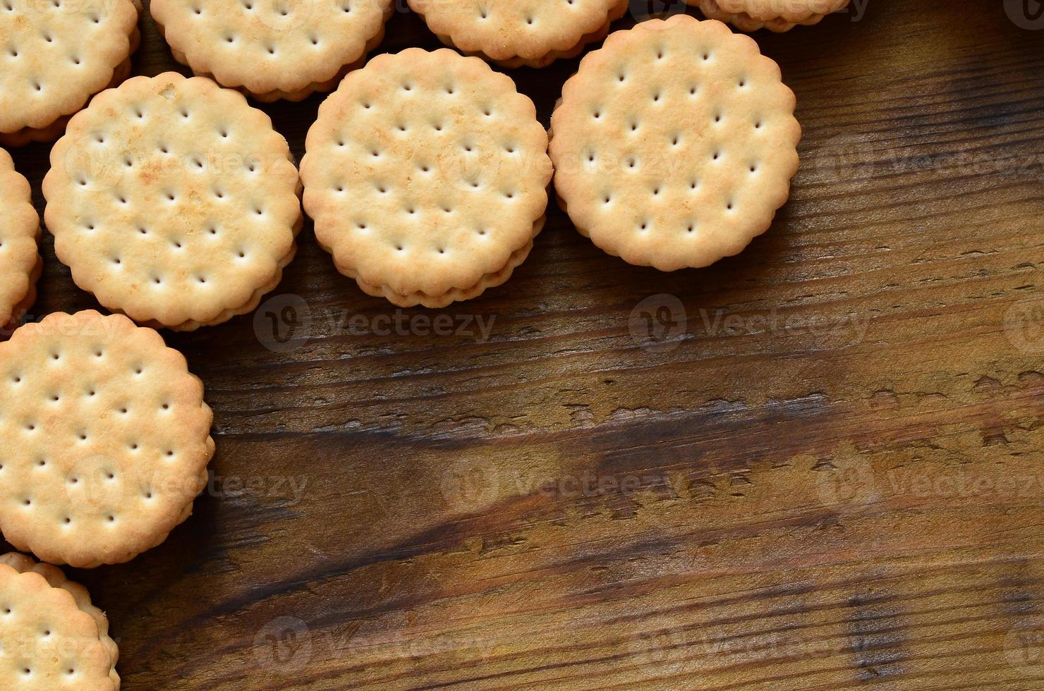 A round sandwich cookie with coconut filling lies in large quantities on a brown wooden surface. Photo of edible treats on a wooden background with copy space