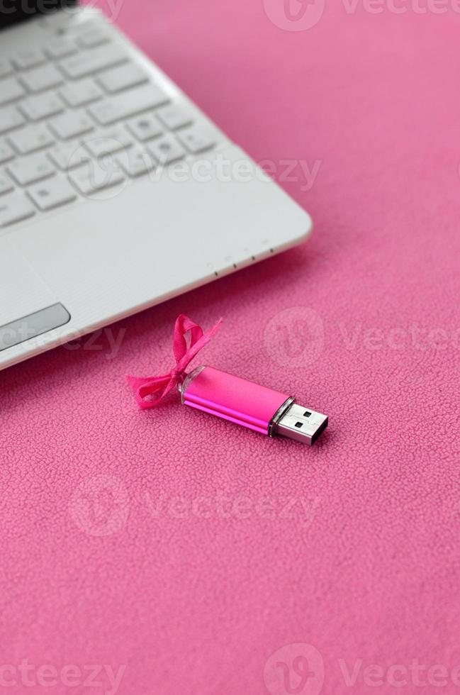 Brilliant pink usb flash memory card with a pink bow lies on a blanket of soft and furry light pink fleece fabric beside to a white laptop. Classic female gift design for a memory card photo