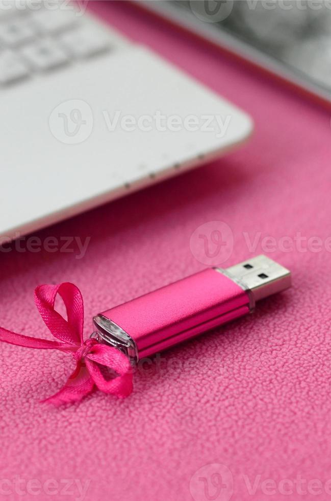 Brilliant pink usb flash memory card with a pink bow lies on a blanket of soft and furry light pink fleece fabric beside to a white laptop and smartphone. Classic female gift design for a memory card photo