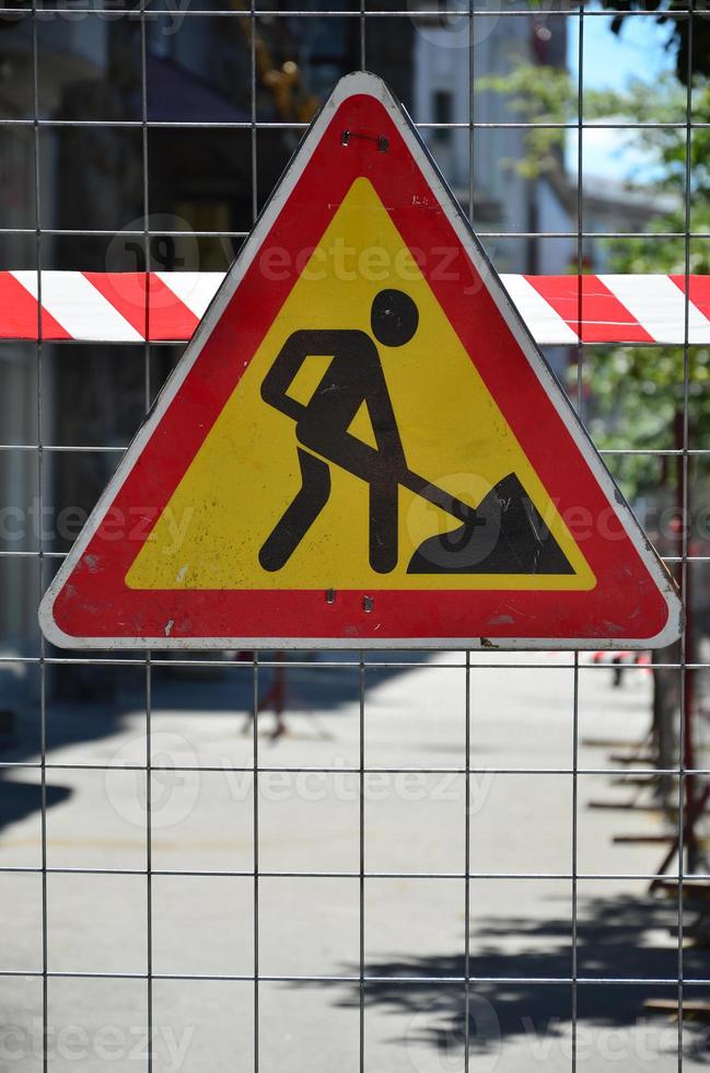 la señal de advertencia en construcción está unida a una cerca de malla metálica con una cinta de señal de rayas rojas y blancas foto