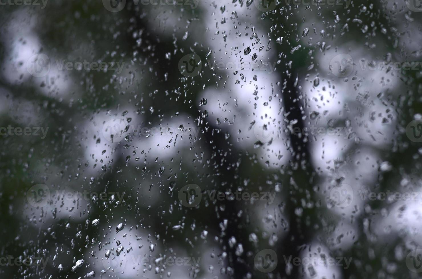 A photo of rain drops on the window glass with a blurred view of the blossoming green trees. Abstract image showing cloudy and rainy weather conditions