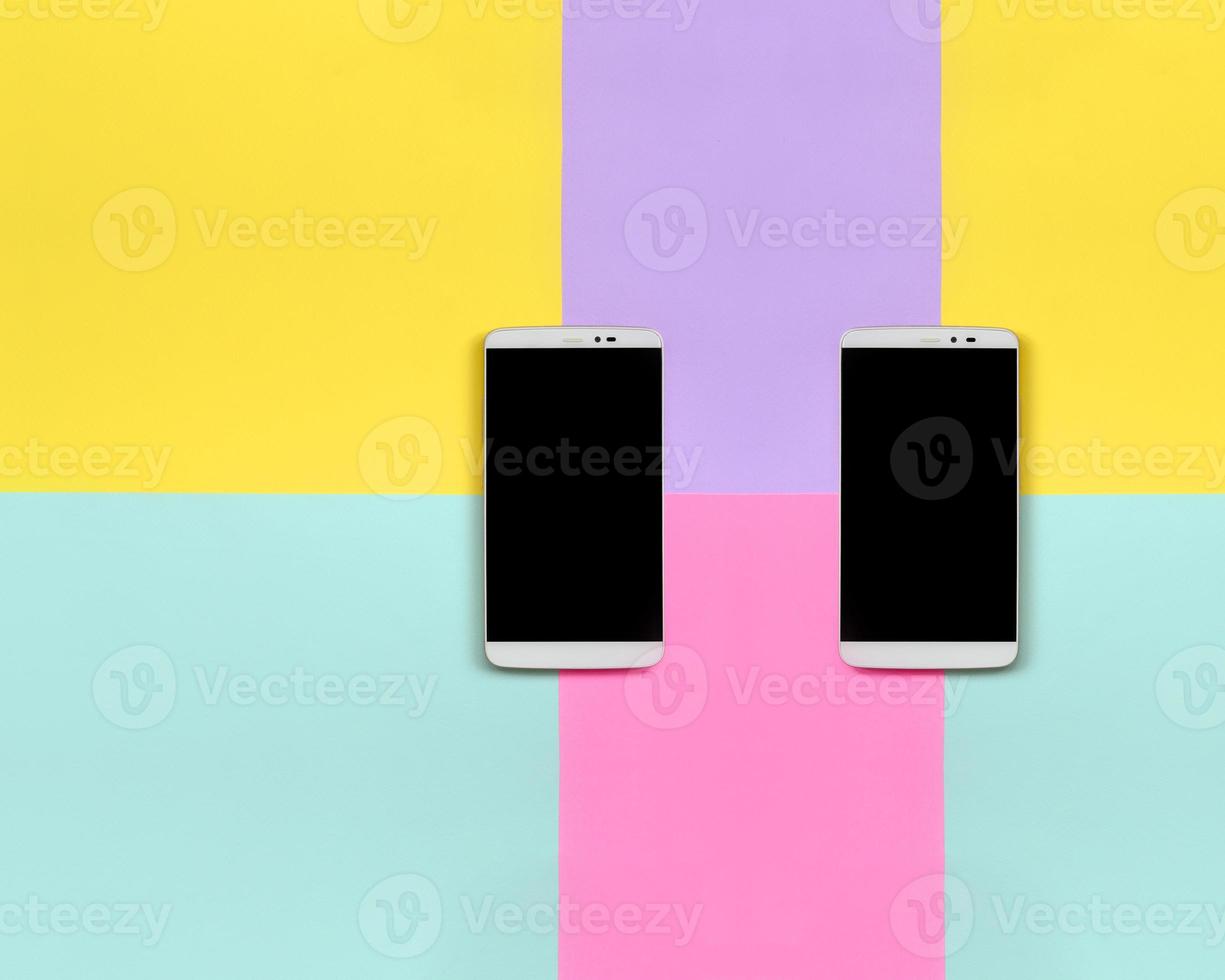Two modern smartphones with black screens on texture background of fashion pastel blue, yellow, violet and pink colors paper in minimal concept photo