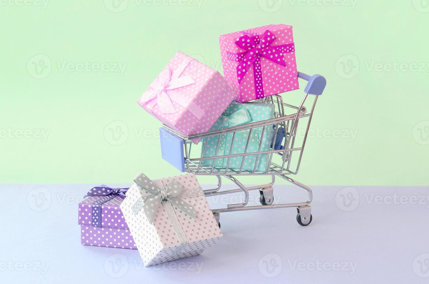 Small gift boxes of different colors with ribbons in shopping cart on a violet and blue pastel background photo