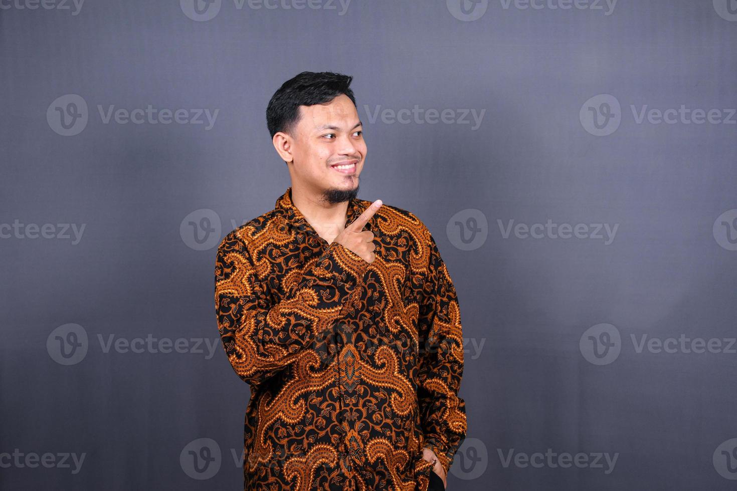 Portrait of a happy young man in batik indonesia pointing fingers away at copy space isolated over gray background photo