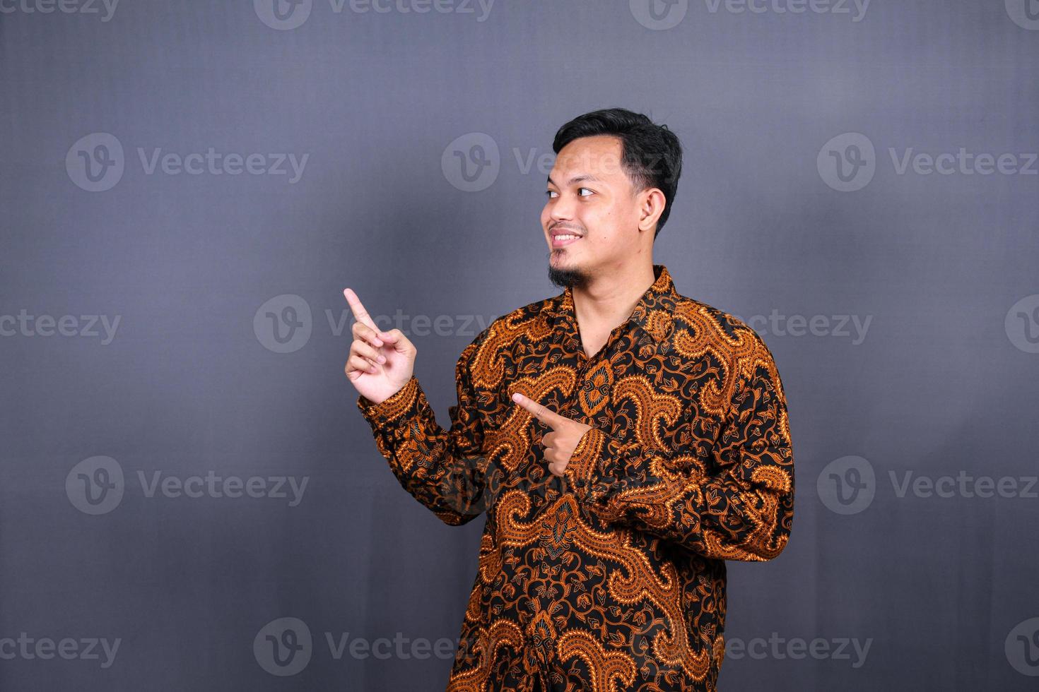 Portrait of a happy young man in batik indonesia pointing fingers away at copy space isolated over gray background photo