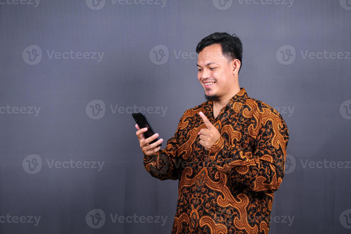 Portrait of asian young man wearing batik using cellphone on gray background photo