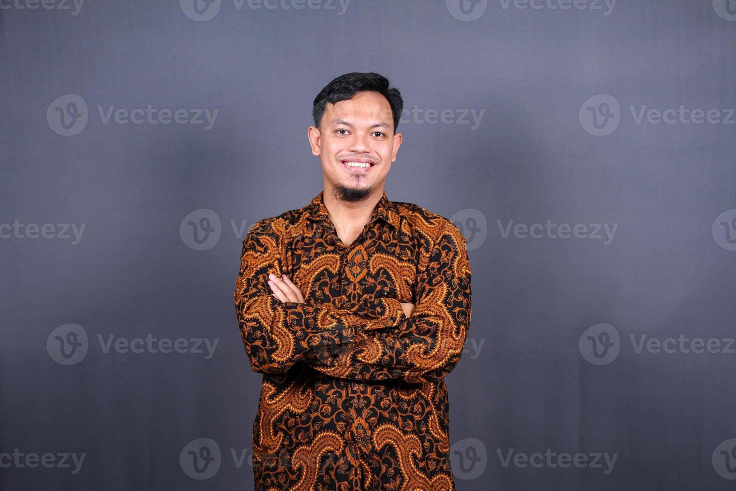 Portrait of attractive young asian man in batik indonesia standing with crossed arms isolated on gray background photo