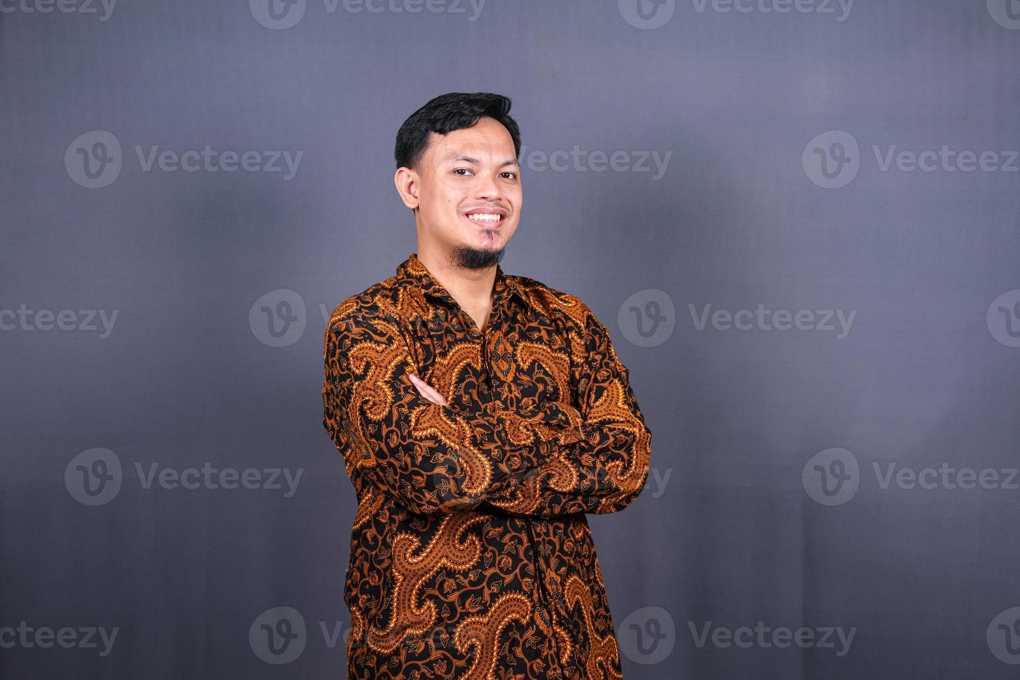 Portrait of attractive young asian man in batik indonesia standing with crossed arms isolated on gray background photo