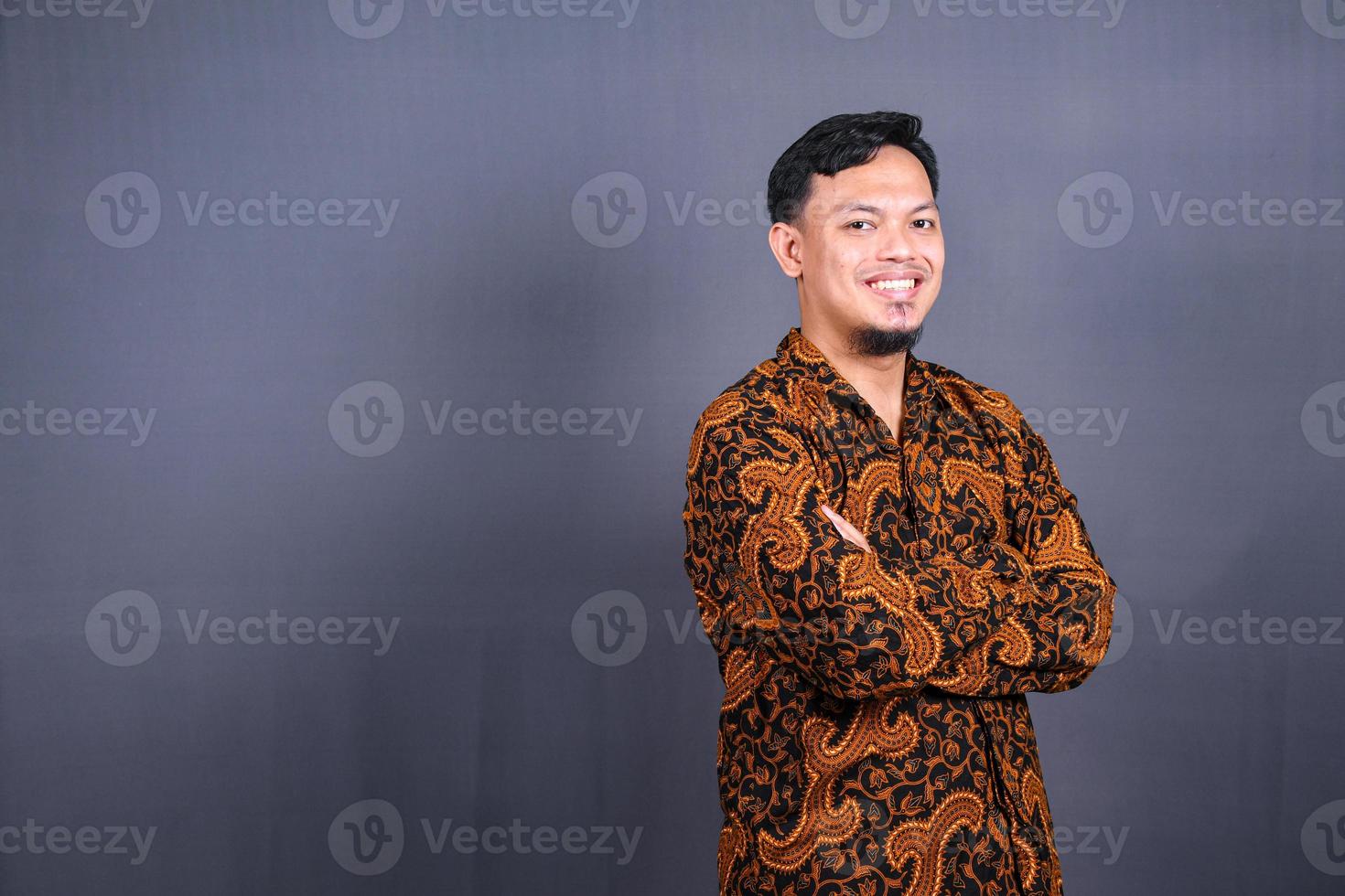 Portrait of attractive young asian man in batik indonesia standing with crossed arms isolated on gray background photo