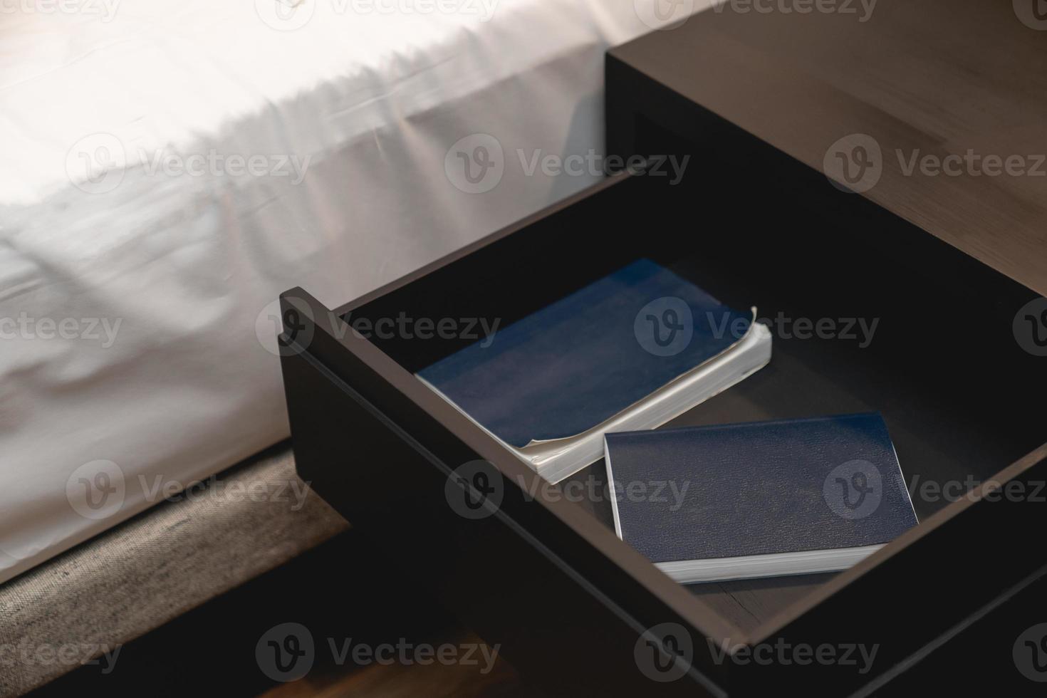 Bed with white sheet and next to wooden drawer stand and note book inside. photo