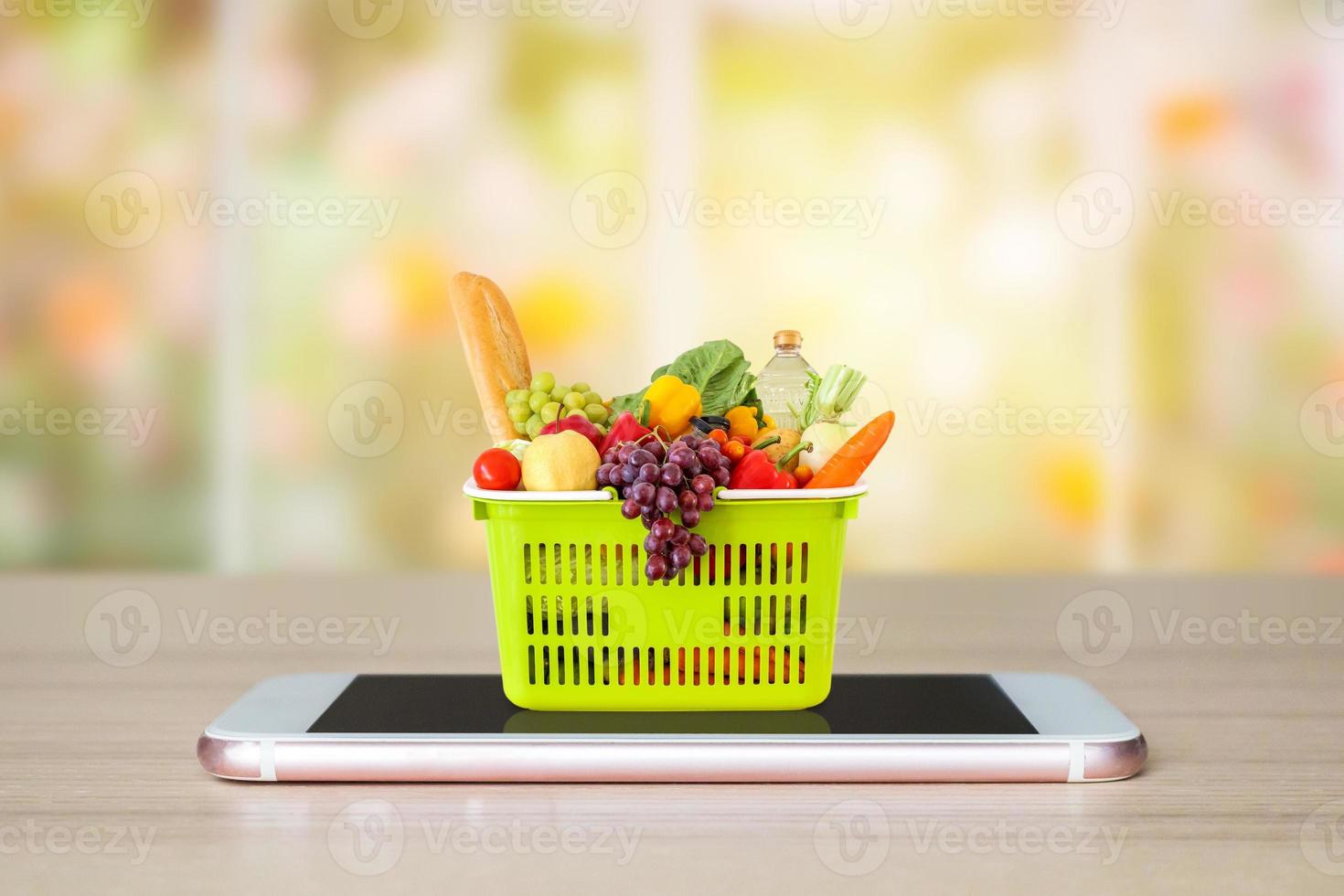 Fresh food and vegetables in green shopping basket on mobile smartphone on wood table with window and garden abstract blur background grocery online concept photo