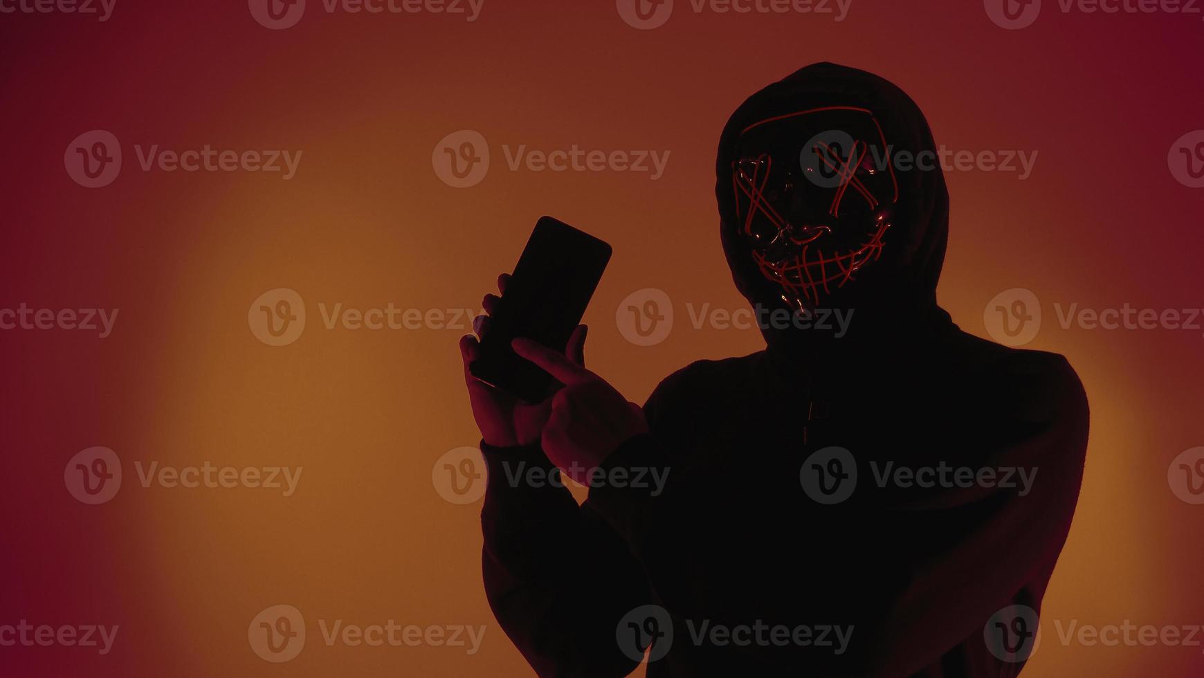 concepto de seguridad digital. hacker anónimo con máscara sosteniendo un teléfono inteligente pirateado. foto