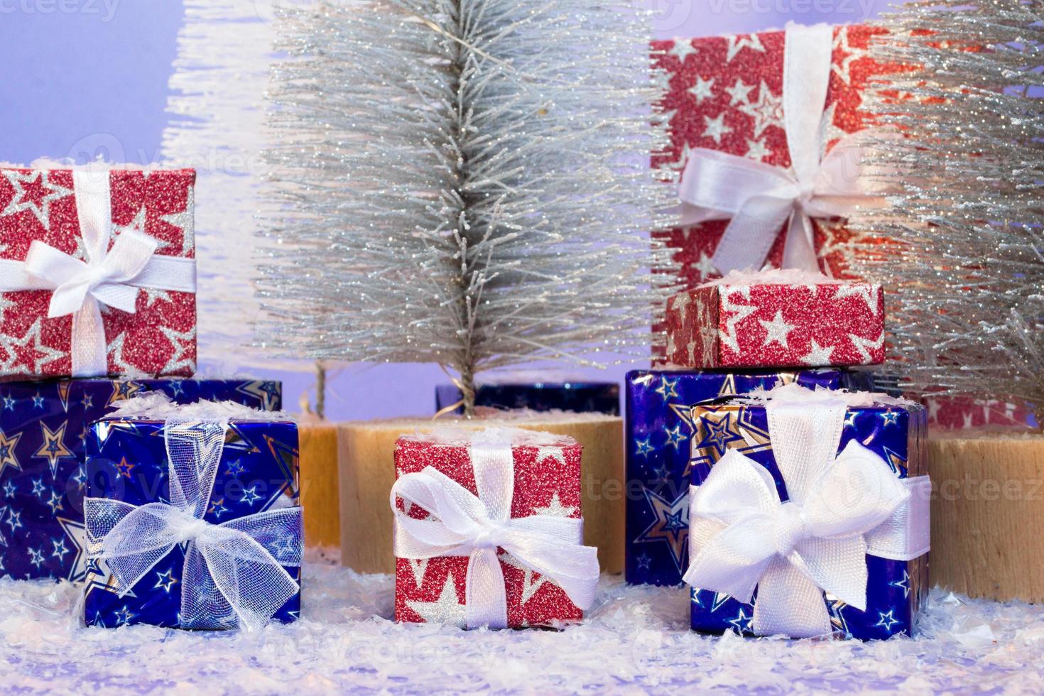 Gift boxes in shiny blue and red paper with artificial snow and Christmas trees on purple background. photo